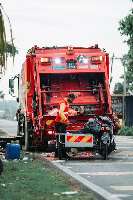 A waste collection worker, dressed in a high-visibility yellow and orange vest, is standing behind the rear of a large red rubbish truck, which is parked on the side of a paved urban street. The worker appears to be attending to the vehicle's compaction or loading mechanism, with their head slightly lowered and hands busy near the equipment. Behind the worker, there is a black plastic garbage bag resting on a metal platform attached to the truck, and various small waste items scattered on the ground nearby. The side of the truck features reflective yellow and red chevron safety markings, and the vehicle displays identification plates and company stickers. In the background, there are trees, power lines, and signage indicating a commercial area, with a partly cloudy sky overhead. This scene exemplifies an independent waste collection or rubbish removal operation in a city environment, aligning with private waste handling services provided by waste clearance companies such as Waste Clearance Chiswick.