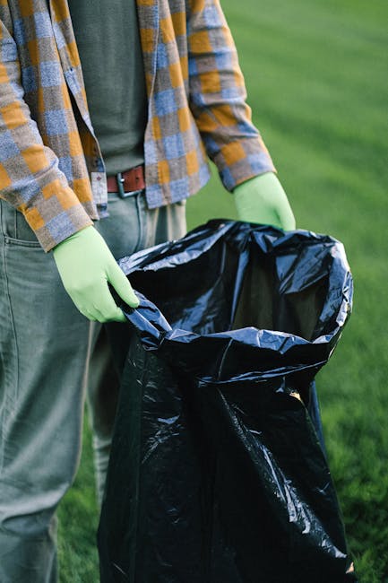 A waste collection worker, dressed in a high-visibility yellow and orange vest, is standing behind the rear of a large red rubbish truck, which is parked on the side of a paved urban street. The worker appears to be attending to the vehicle's compaction or loading mechanism, with their head slightly lowered and hands busy near the equipment. Behind the worker, there is a black plastic garbage bag resting on a metal platform attached to the truck, and various small waste items scattered on the ground nearby. The side of the truck features reflective yellow and red chevron safety markings, and the vehicle displays identification plates and company stickers. In the background, there are trees, power lines, and signage indicating a commercial area, with a partly cloudy sky overhead. This scene exemplifies an independent waste collection or rubbish removal operation in a city environment, aligning with private waste handling services provided by waste clearance companies such as Waste Clearance Chiswick.