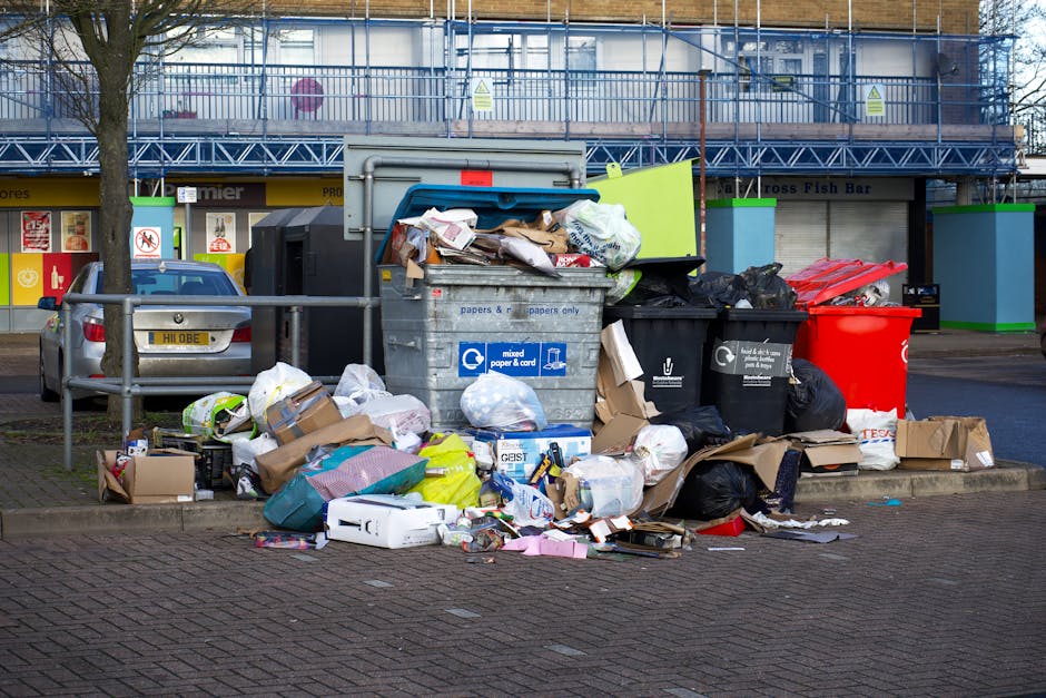 A large collection of rubbish, including black plastic trash bags, brown paper bags, cardboard boxes, and loose paper, is piled on a paved area beside a metal railing near a parking lot in an urban environment. A grey plastic bin with a blue lid, labeled for mixed paper and cardboard recycling, is partially open with various waste materials spilling out onto the ground. To the right, there is a black bin and a red bin, both filled with waste, with some contents overflowing and scattered around the base. In the background, there are commercial storefronts with signage, a parked grey car on the left, and a building with scaffolding or construction barriers on the upper level, indicating an area with disposal or waste clearance activity. Natural light illuminates the scene, highlighting the textures and colors of the assorted waste materials, visually representative of private rubbish collection or on-site clearance typical for independent waste handling solutions like those offered by waste clearance services.