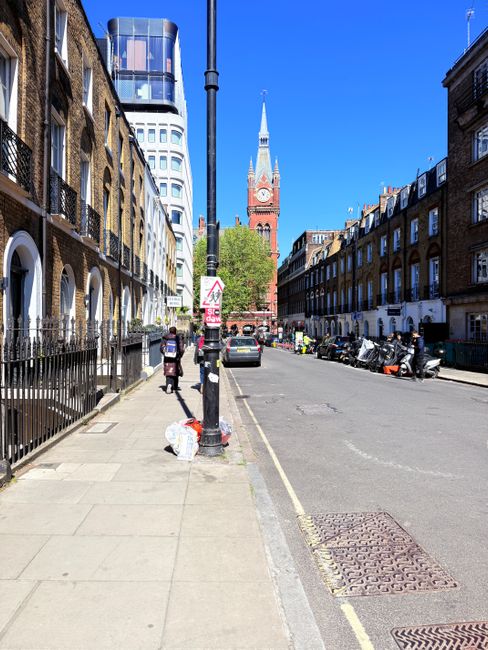 A view of an urban street in daylight showing a sidewalk with a black metal railing and two pedestrians walking, one carrying a small bag, beside a row of brick residential buildings with arched windows and white detailing. A black lamp post is positioned centrally on the sidewalk with a plastic bag and a small pile of rubbish placed nearby. On the street, several parked motorcycles and small cars are visible, along with a few people near the vehicles. In the background, a tall clock tower with a pointed spire and a clock face is prominent against a clear blue sky. Surrounding the tower are modern and historic buildings, including one with distinctive rounded glass architecture. The scene conveys a typical city street environment with a mix of old and new architecture, and the setting suggests areas where private rubbish removal or on-site clearance services, such as those offered by Waste Clearance Chiswick, might be engaged for waste management needs.