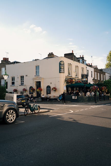 A street scene in front of a white, two-story building with a traditional pub facade, featuring large windows, flower baskets hanging from the exterior walls, and a green canopy extending over an outdoor seating area. The sidewalk is occupied by pedestrians and patrons seated at tables, while a black vehicle and a bicycle are parked on the road in the foreground. The surrounding environment includes a clear blue sky with some scattered clouds, and additional historic-style buildings lining the street, suggesting an urban area that may be serviced by private waste collection or alternative rubbish removal options such as waste clearance services like Waste Clearance Chiswick, located in close proximity to shops, restaurants, and public areas.