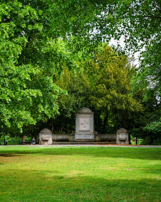 A stone monument situated in the middle of a lush, green park with tall trees providing a canopy of bright green leaves. The monument features a rectangular central structure with a carved relief, flanked by two smaller, curved stone benches on either side. The foreground consists of a well-maintained grassy lawn, while the background shows dense foliage and trees that frame the scene. The lighting is natural, suggesting a cloudy day with diffuse light evenly illuminating the area. The setting and visible elements imply a peaceful outdoor space that could be associated with private or independent preservation efforts, typical of a park or historic site where rubbish removal or site clearance services might be necessary for maintenance or restoration work, as offered by companies like Waste Clearance Chiswick.