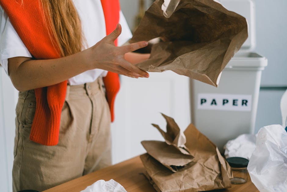 The image shows a person wearing a white shirt and khaki trousers, visible from the waist down, standing in front of a white paper recycling bin labeled 'PAPER'. The individual is holding a crumpled brown paper bag with both hands, preparing to dispose of it. On the wooden table in front of them, there are additional crumpled paper bags and some white plastic waste, indicating active waste separation or refuse collection. The background is a clean, indoor environment with soft, diffused lighting, highlighting the textures of the paper and the smooth surface of the table. This scene exemplifies organized waste management practices related to rubbish removal, specifically emphasizing the separation of paper waste for appropriate disposal or recycling, fitting within the context of private rubbish clearance services offered by companies like Waste Clearance Chiswick, which facilitate on-site waste handling and disposal options outside of standard local authority collections.